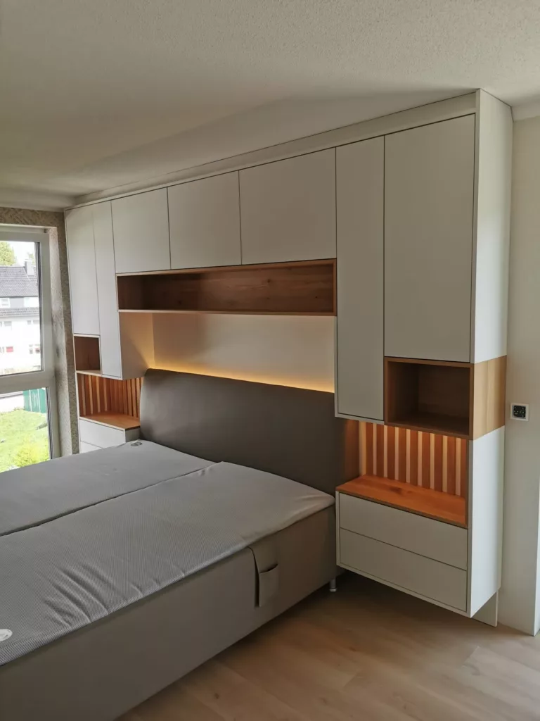 Schlafzimmer mit Bettüberbau in Weiß matt, Nischen und Lamellen aus Eiche und indirektem Licht in Korbach